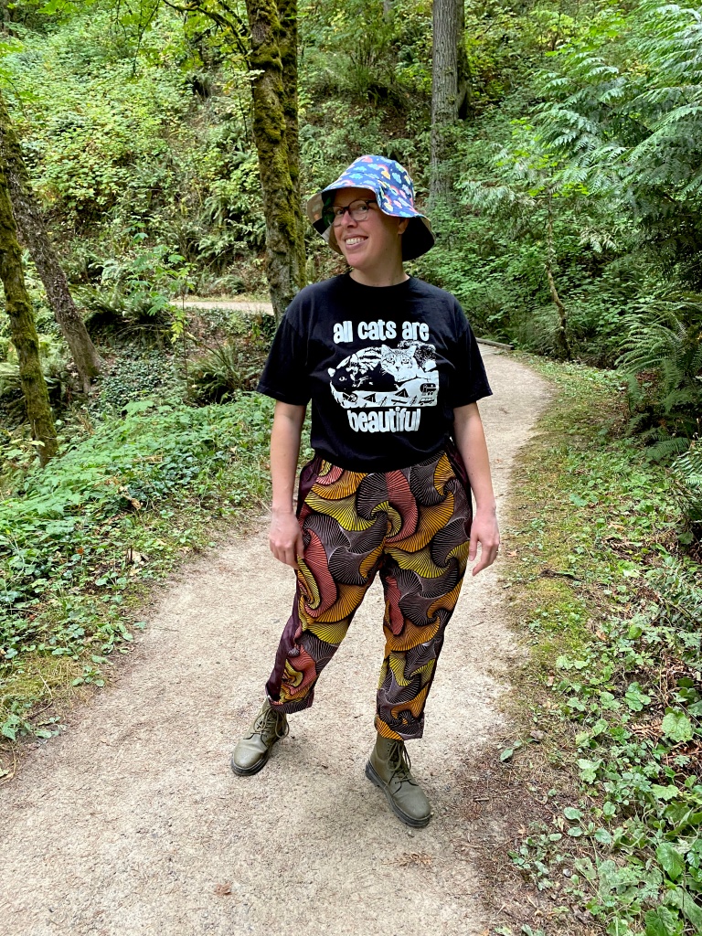 Lee wearing colorful pants, a tee that says All Cats Are Beautiful, and a bucket hat, on a trail in the forest