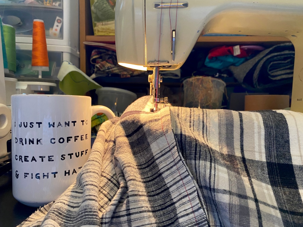 a vintage sewing machine with flannel fabric going through it, and a mug sitting next to it that says "I just want to drink coffee, create stuff & fight hate"
