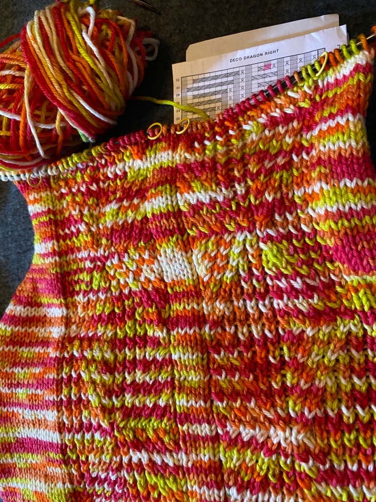 an in-progress knit in orange, pink, white, and green, with a textured twisted stitch pattern