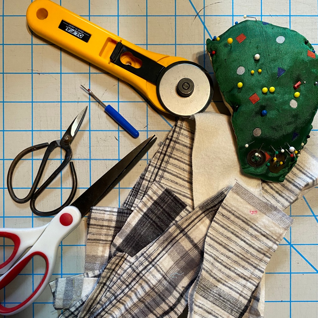 top-down photo of a cutting mat with some flannel fabric strips, 2 pairs of scissors, a seam ripper, a rotary cutter, and a pin cushion