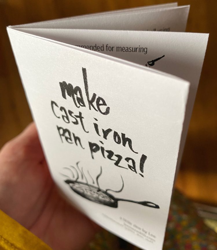 hand holding folded zine with cover saying "make cast iron pan pizza!" in handlettering, with a drawing of a pan with stream lines coming up from it