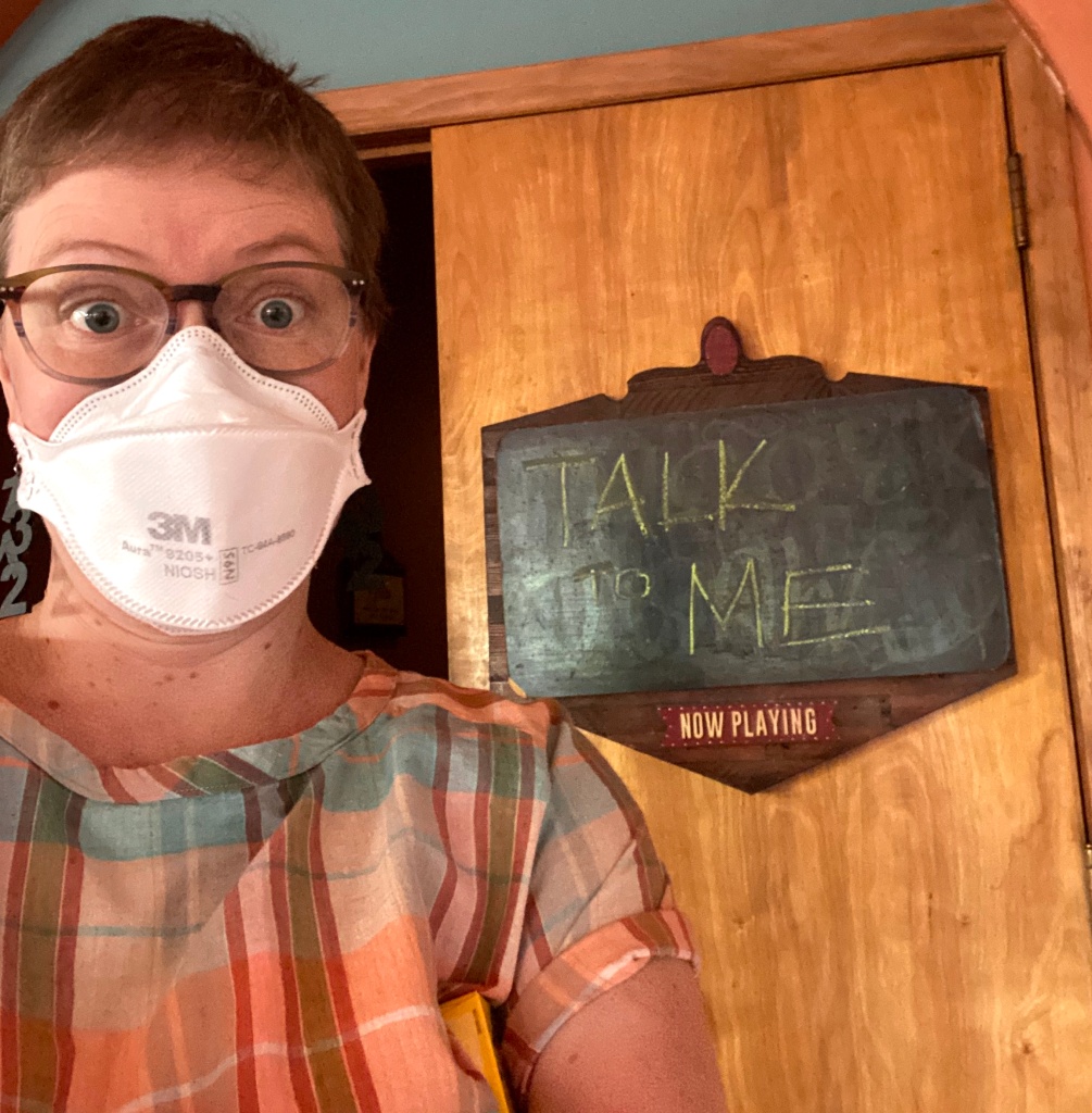 Lee wearing a plaid t-shirt and a mask standing in front of a wooden door with a chalkboard sign that says Talk to Me now playing