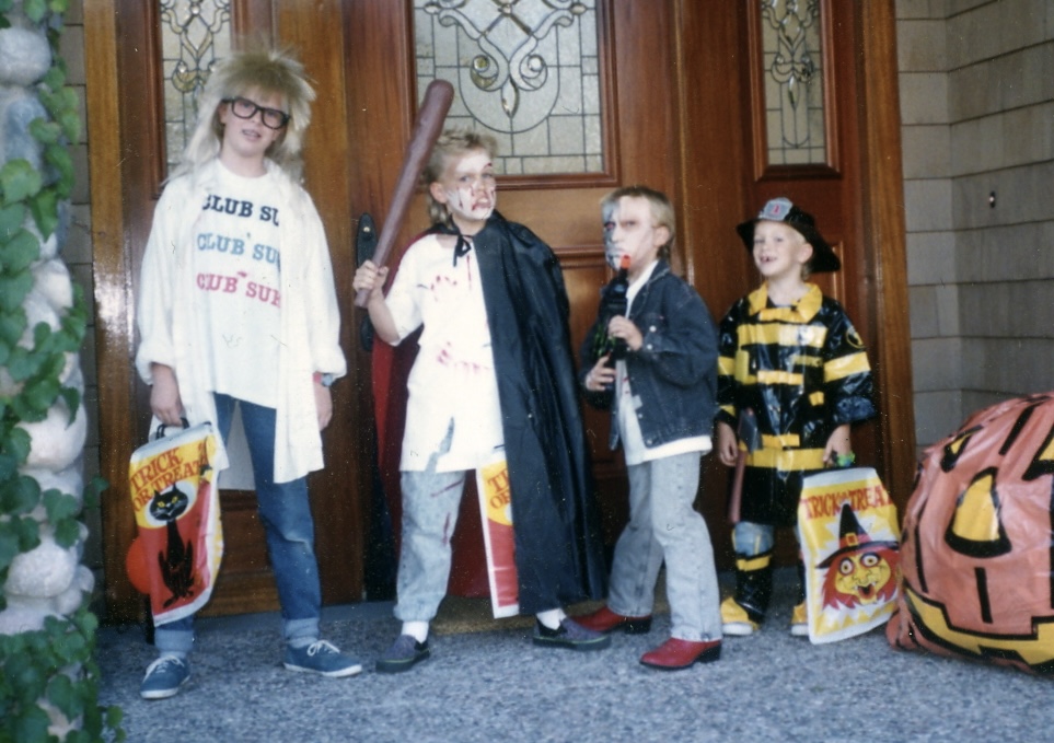 old photo of four kids dressed up for Halloween, lined up tallest to shortest in front of a door. first is dressed as Garth from Wayne's World, second is some kind of blood-splattered bad guy with a bat-like weapon, third is wearing a mask with metal on one half and a black denim jacket and red boots, and the fourth is a firefighter