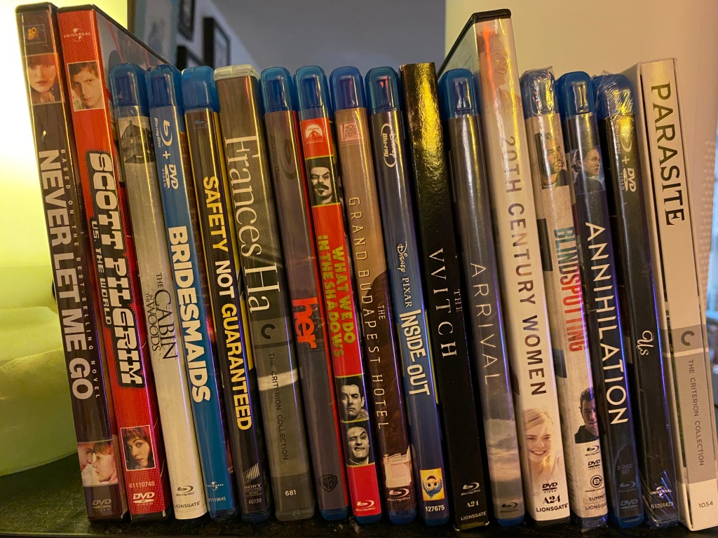 DVD or blu ray copies of these movies lined up on a shelf: never let me go, scott pilgrim, cabin in the woods, bridesmaids, safety not guaranteed, frances ha, her, what we do in the shadows, grand budapest hotel, inside out, the witch, arrival, 20th century women, blindspotting, annihilation, us, parasite